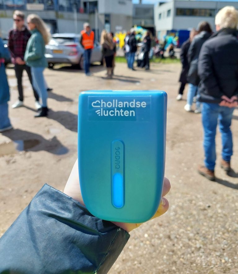 Iemand die een Hollandse Luchten sensor vasthoudt. Op de achtergrond staan mensen met elkaar te praten.