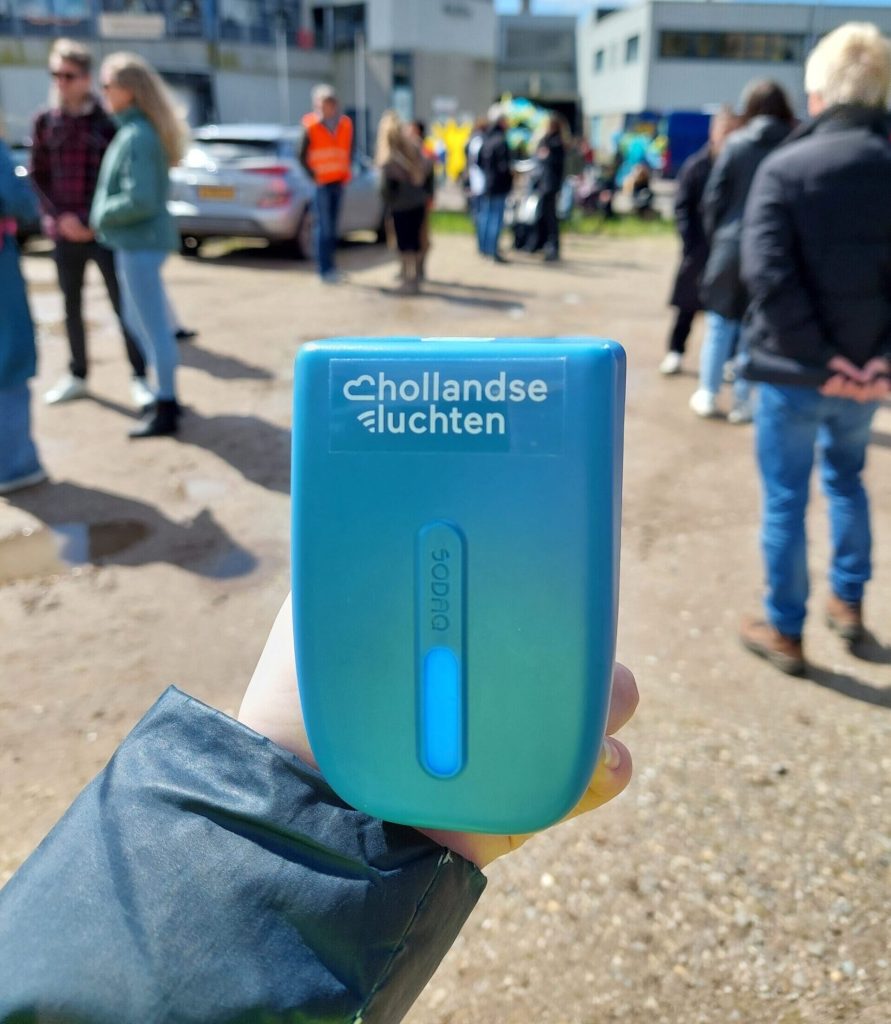 Iemand die een Hollandse Luchten sensor vasthoudt. Op de achtergrond staan mensen met elkaar te praten.